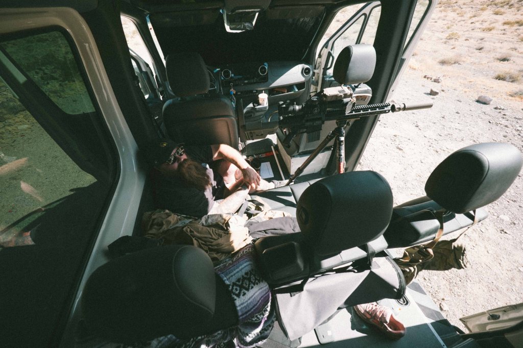 Interior view of a vehicle with two individuals resting; one is lying on the floor while the other sits nearby. There is a mounted firearm visible on the seat, and the environment appears rugged with dirt and sparse vegetation outside.